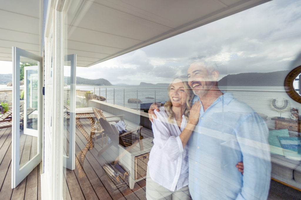 Mature couple looking at the view in their waterfront home. They look happy and contented. They are embracing looking through a window. The ocean can be seen in the reflection