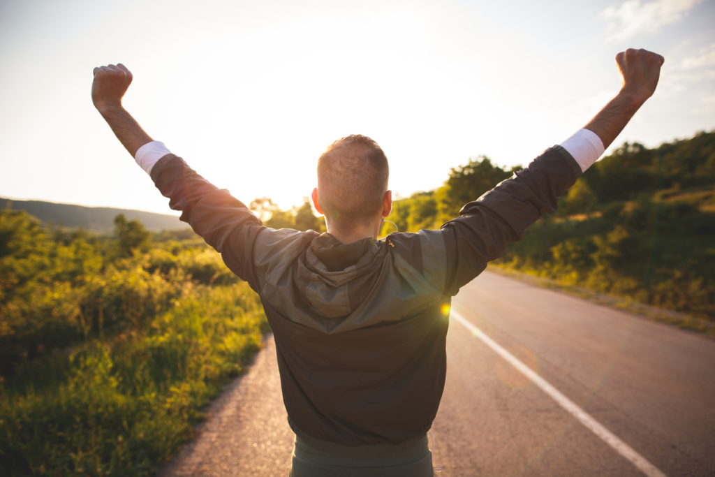 Sport and life achievements and success concept. Sporty man raising arms towards beautiful sunset on the road