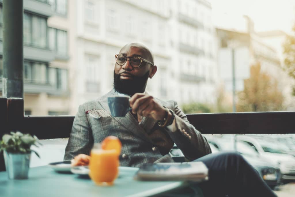 The portrait of a handsome stylish wealthy African guy with a beautiful black beard, in glasses, bald, in an elegant suit, sitting on a rainy morning in a street cafe and drinking delicious coffee